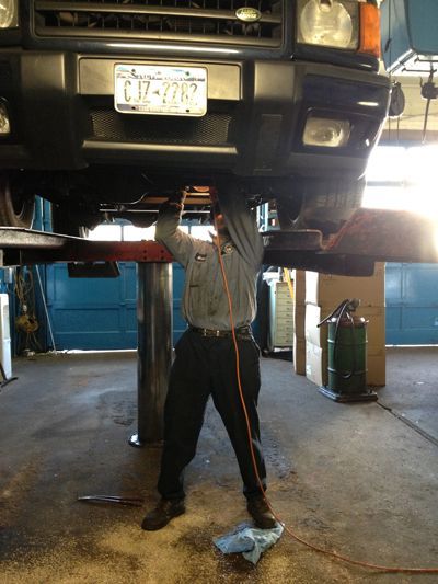 Mechanic working under a raised car in a garage; wearing gray shirt and black pants.