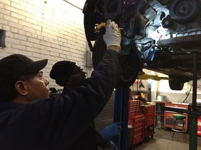 Two mechanics working on a car lifted on a hoist in a garage. One wears gloves, the other a beanie.