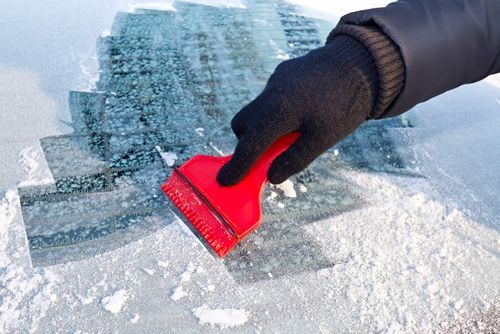 Gloved hand scraping ice from a car windshield with a red scraper.