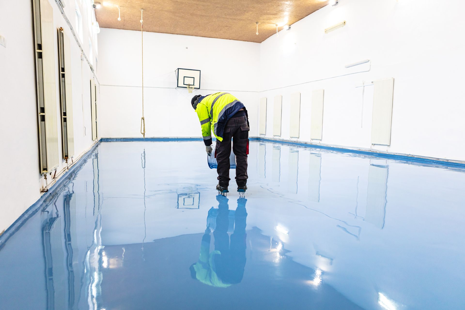 Worker in a high-visibility jacket inspecting a glossy blue floor in a white industrial room