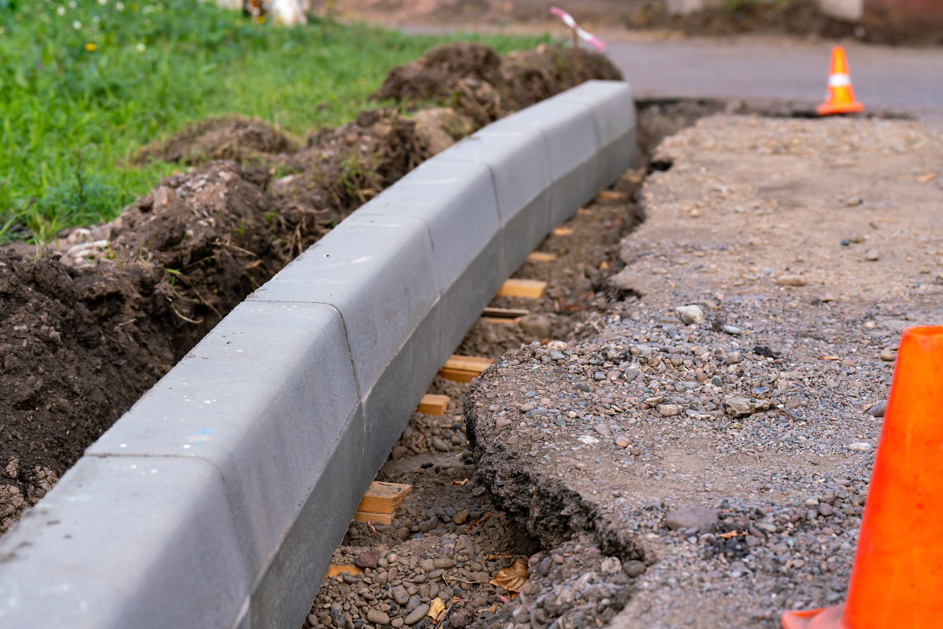 Damaged curb beside a broken road edge with orange cones and dirt excavation.