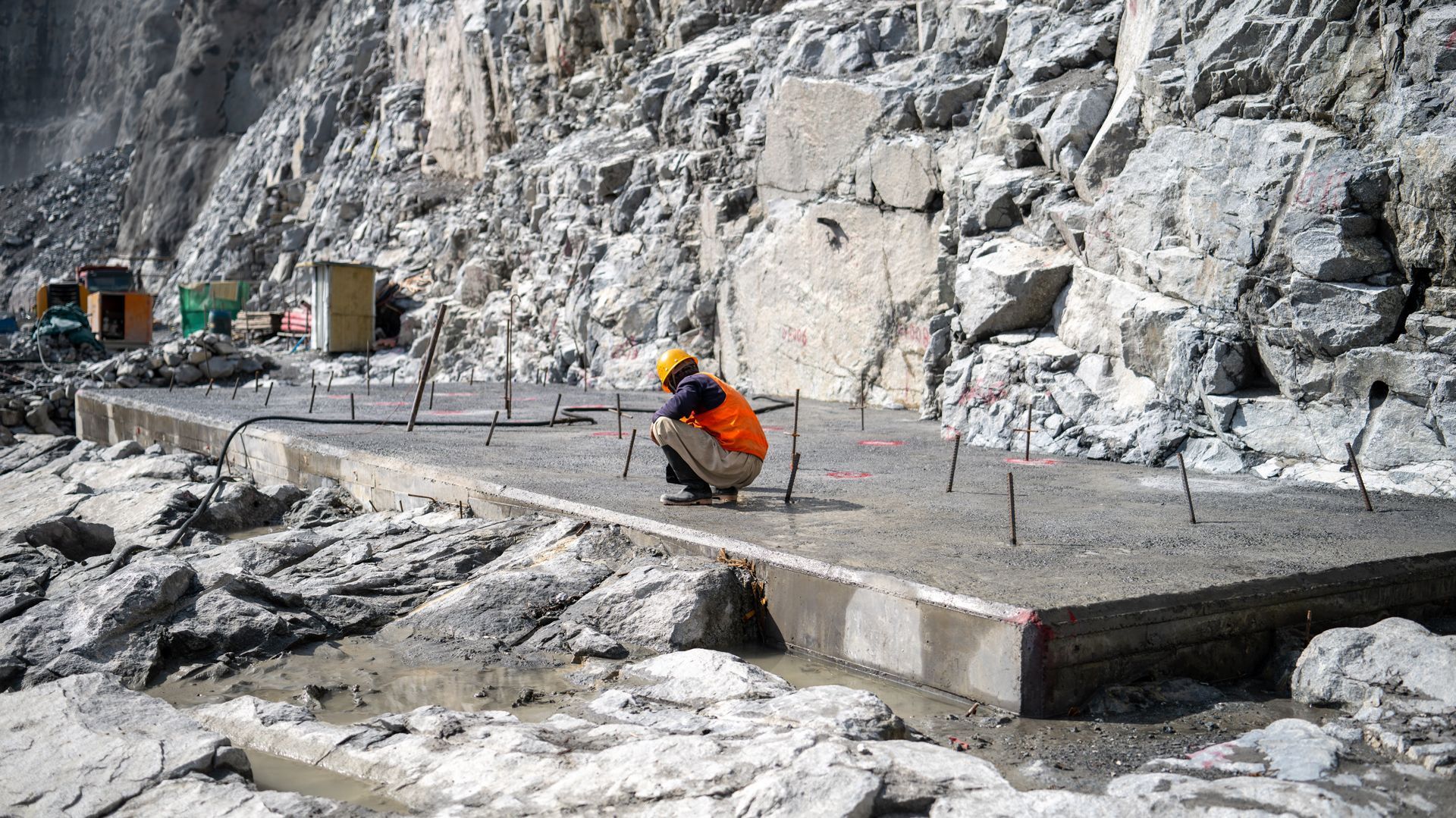 Worker in orange vest crouches on a concrete platform at a rocky cliffside construction site.