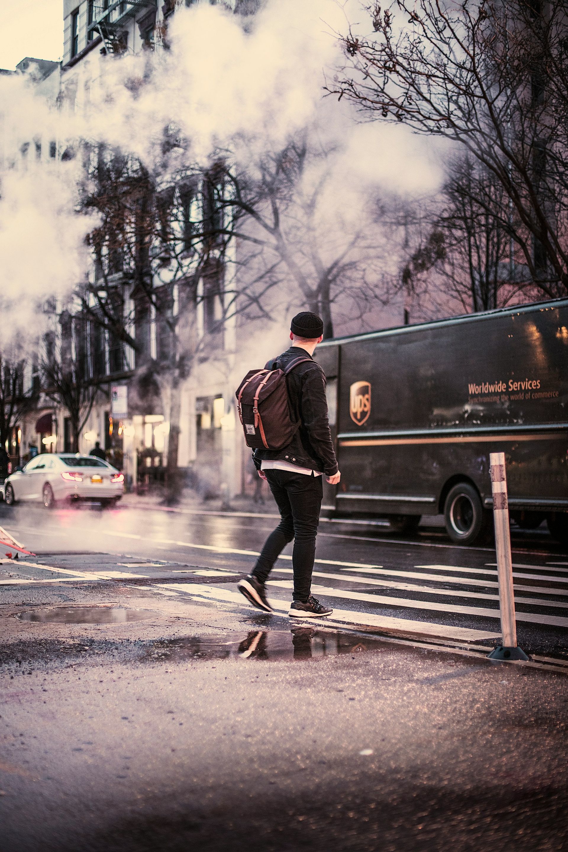 A man with a backpack is crossing the street in the rain.