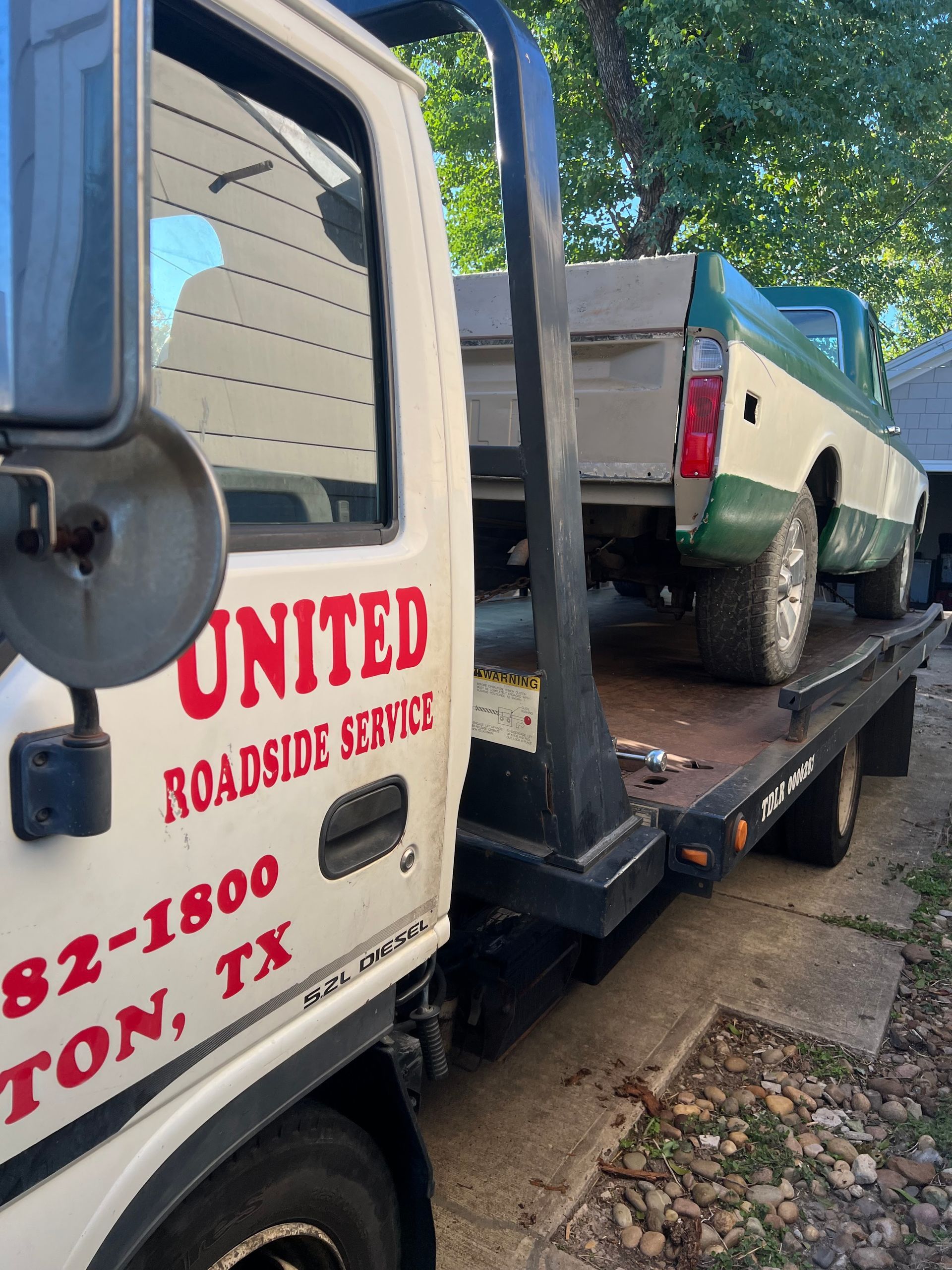 Tow truck hauling a white pickup on a flatbed, parked by a curb under trees