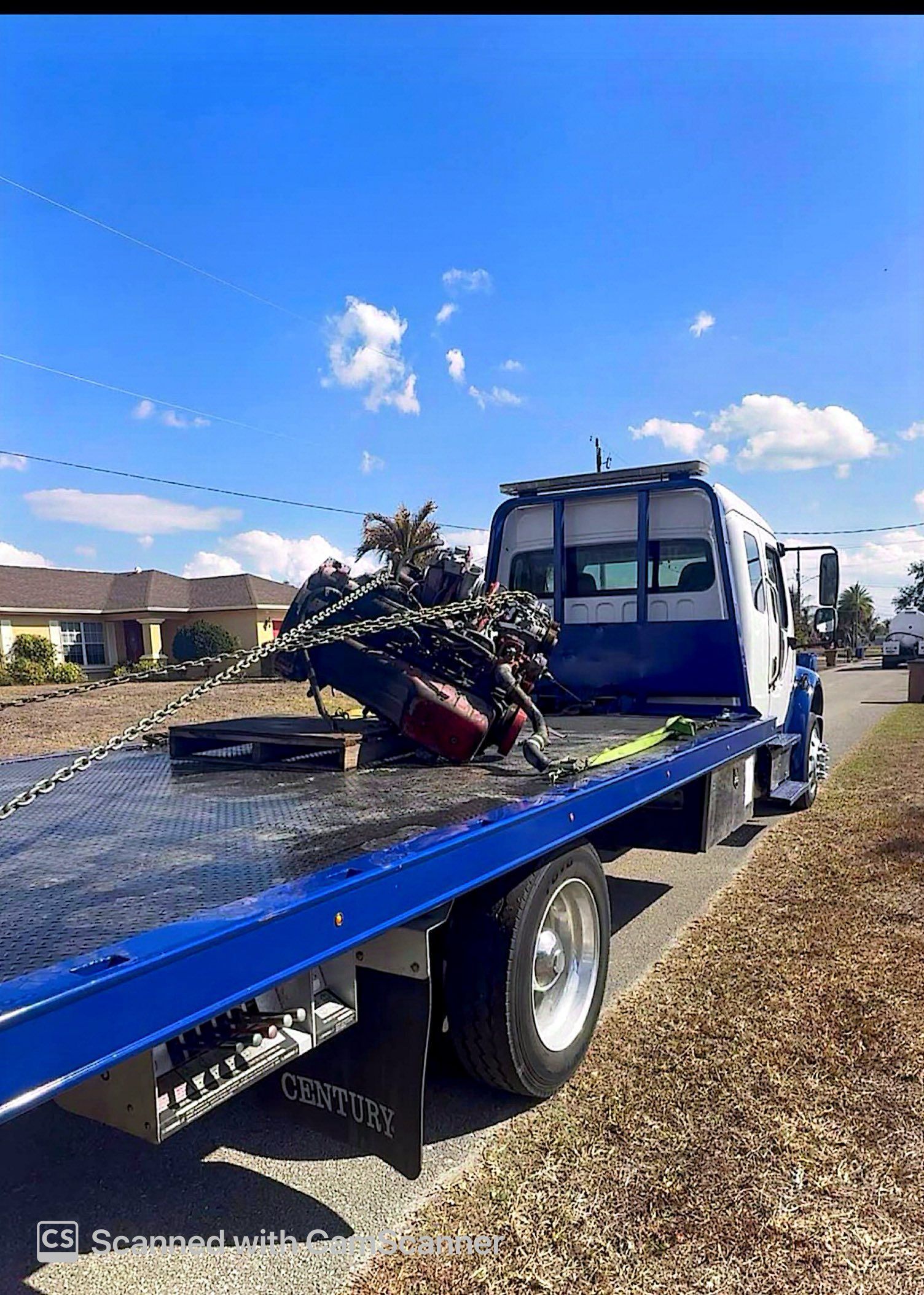 Blue flatbed tow truck carrying a wrecked car on a gravel road under a clear sky