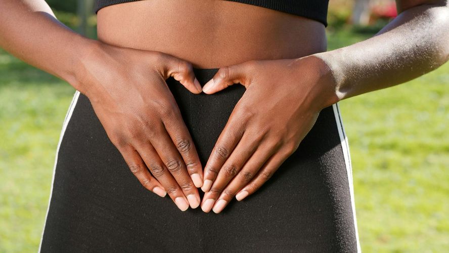 Hands form a heart shape over a person's abdomen, outdoors with greenery.