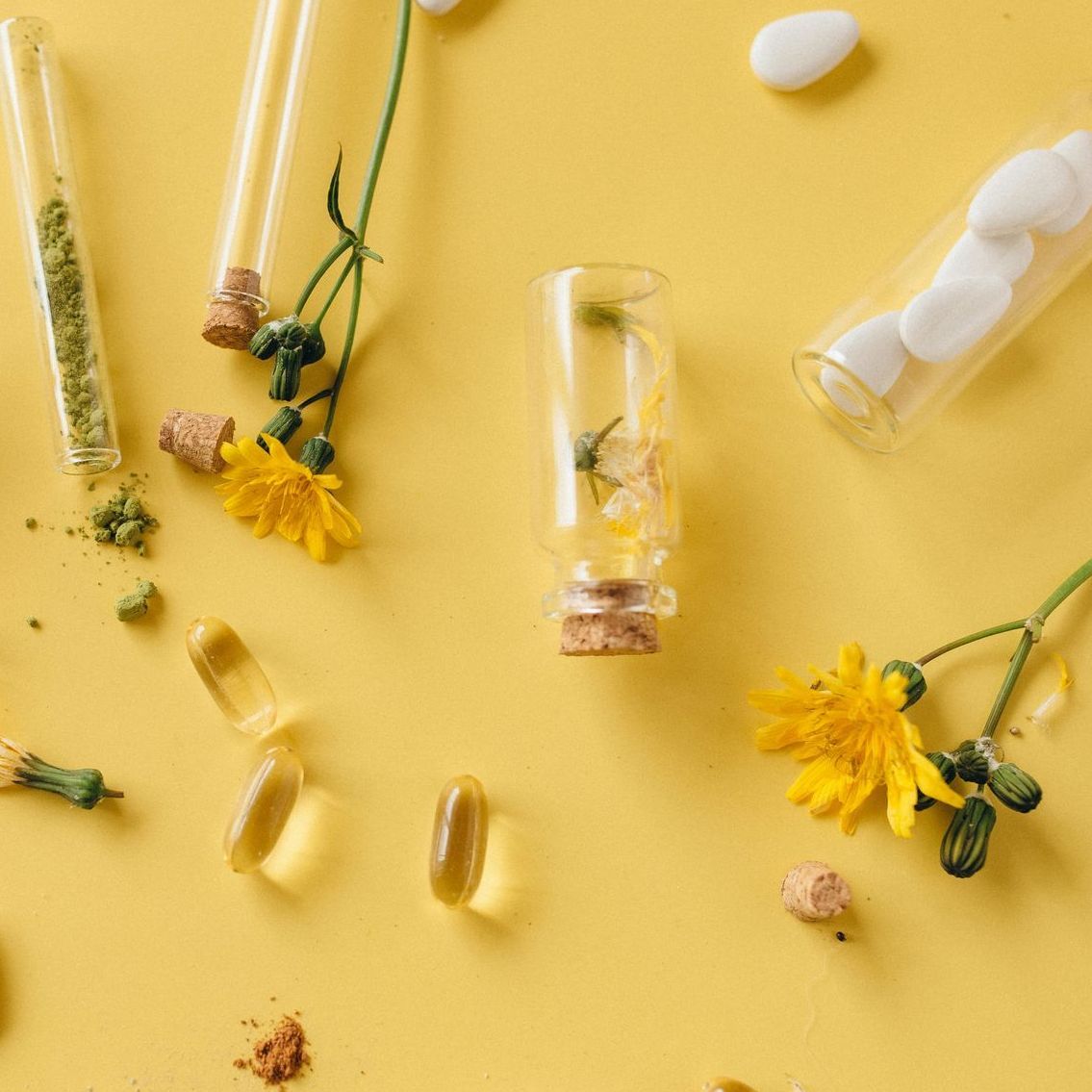 Test tubes with herbs, pills, and flowers on a yellow surface.