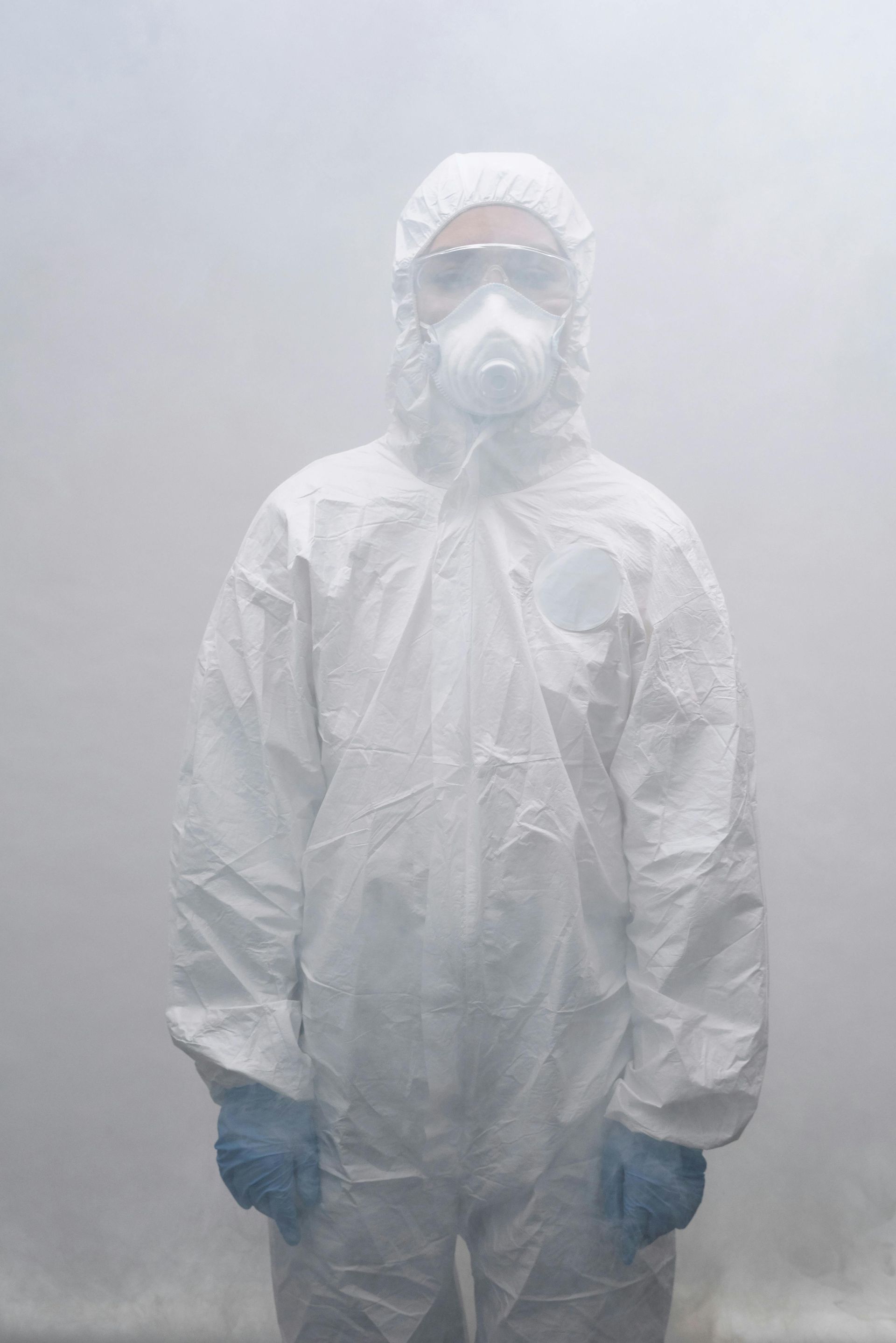 Person in a white hazmat suit, respirator, and gloves, standing in a foggy environment.