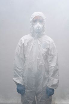 Person in a white hazmat suit, respirator, and gloves, standing in a foggy environment.