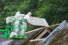 Two workers in protective suits removing asbestos roof tiles from a building, using a lift.