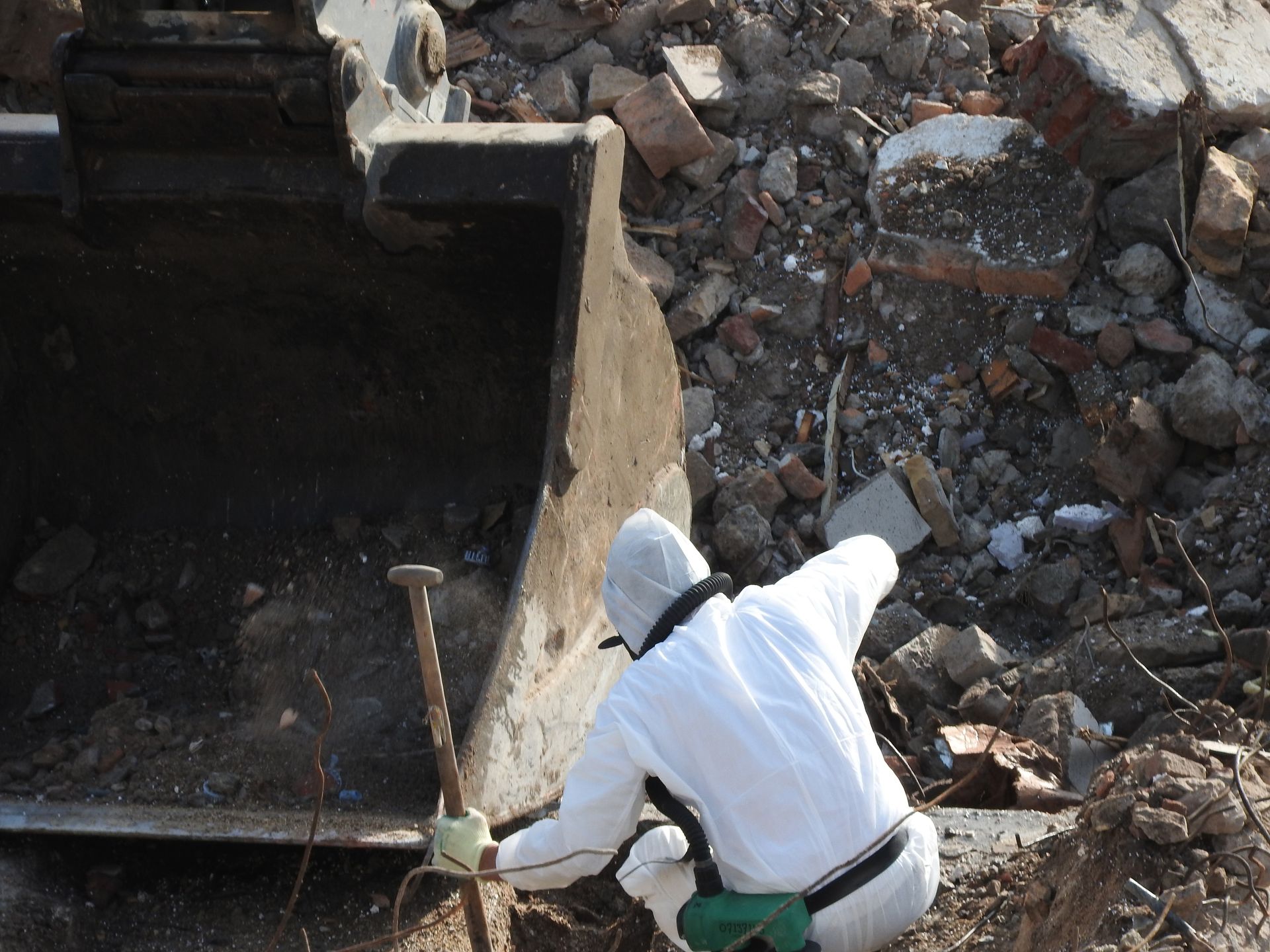 Person in white hazmat suit examining rubble near an excavator bucket.