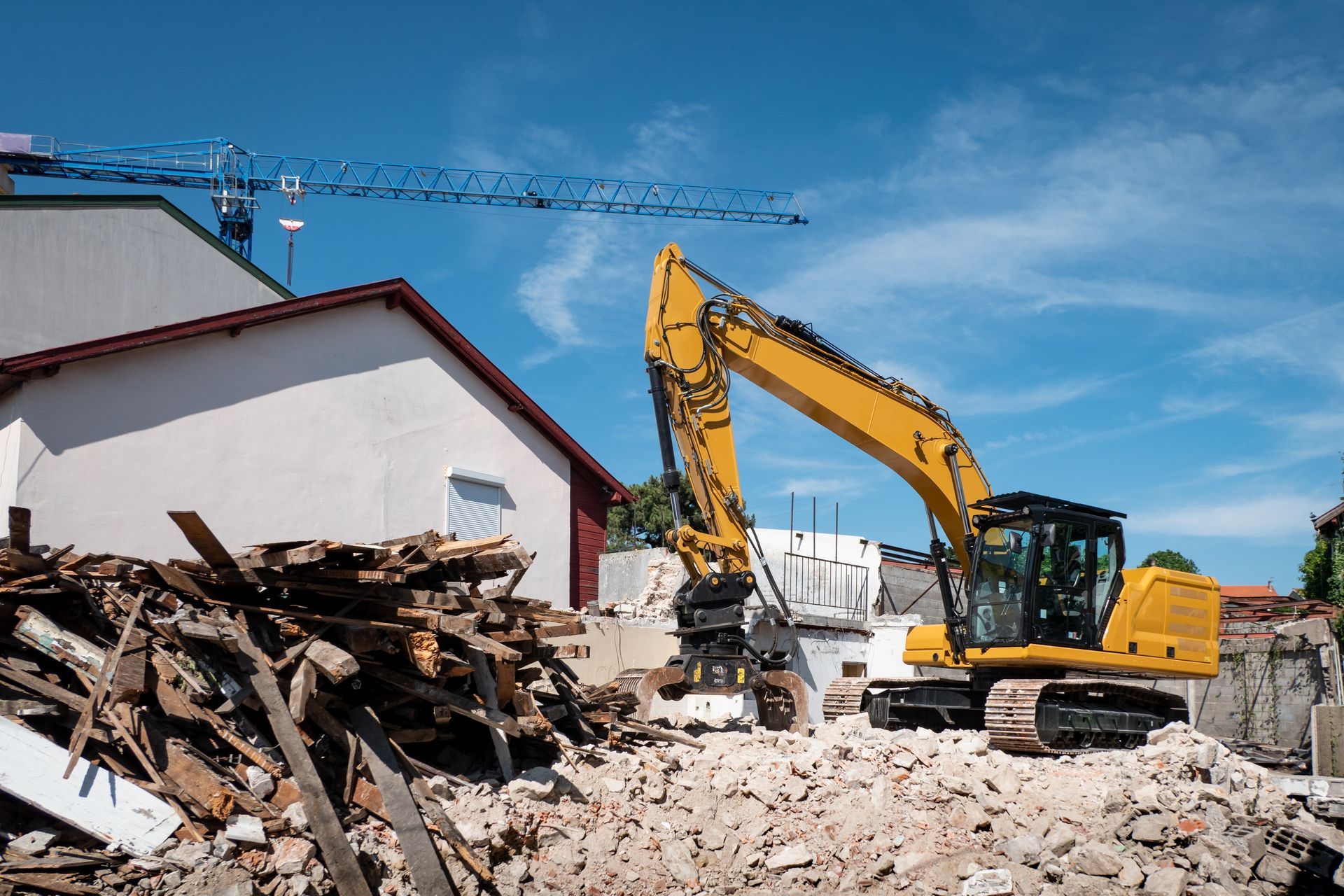 Yellow excavator demolishing a building; pile of debris, blue sky, construction crane in the background.