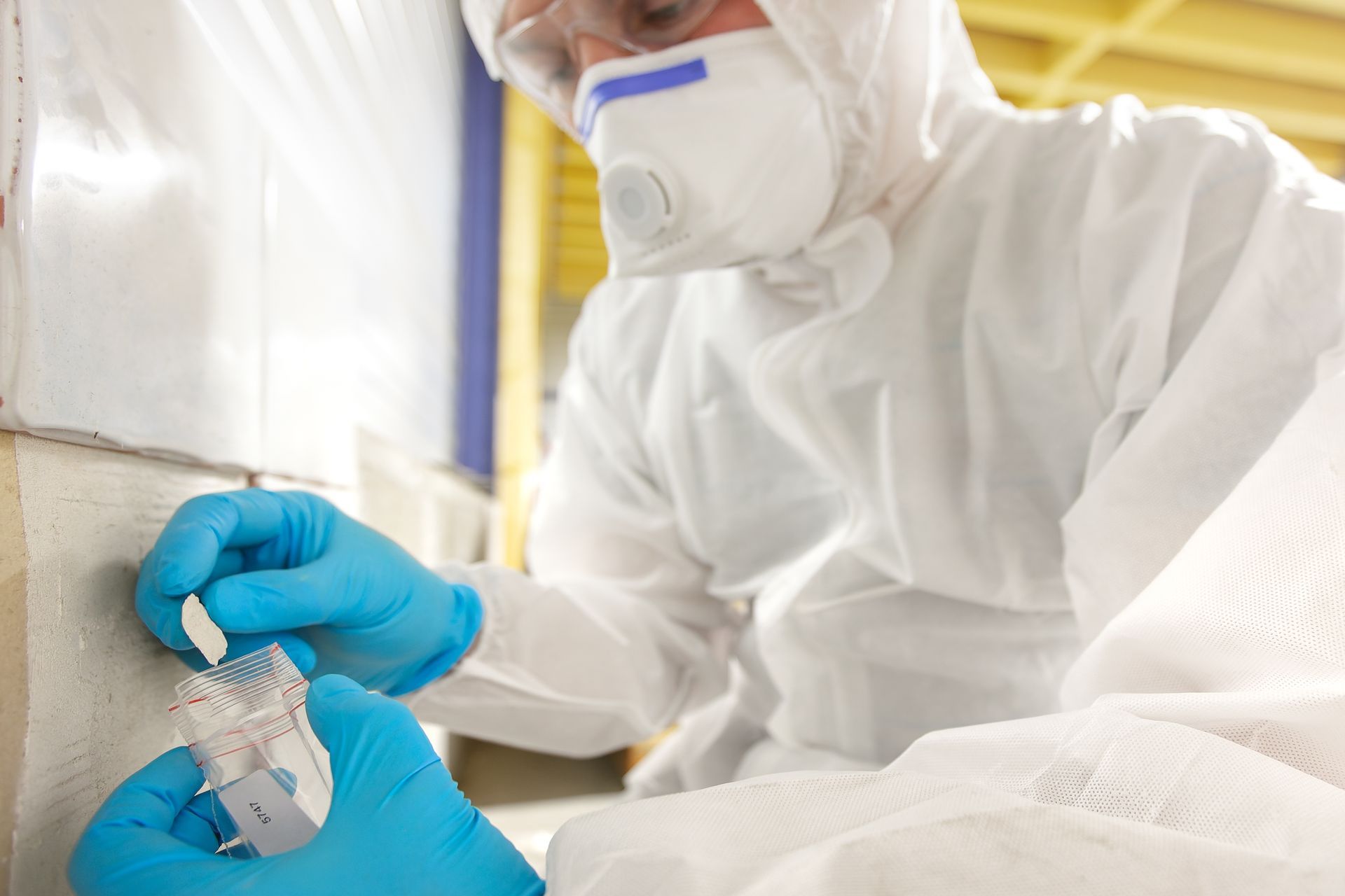 Person in hazmat suit and gloves examines an object near a wall; protective gear and laboratory setting.