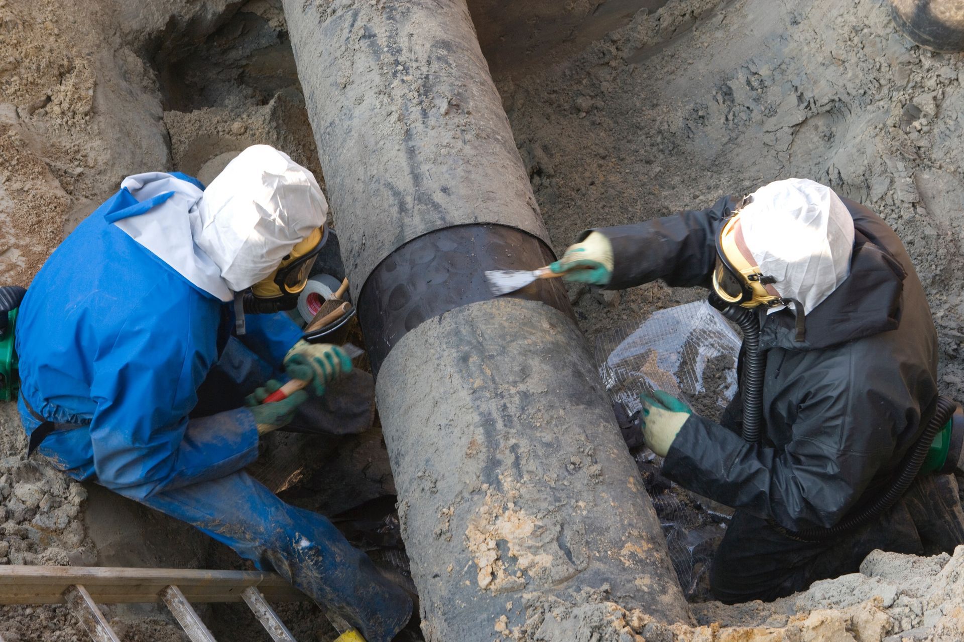 Two workers in protective suits repairing a large pipe in a trench.