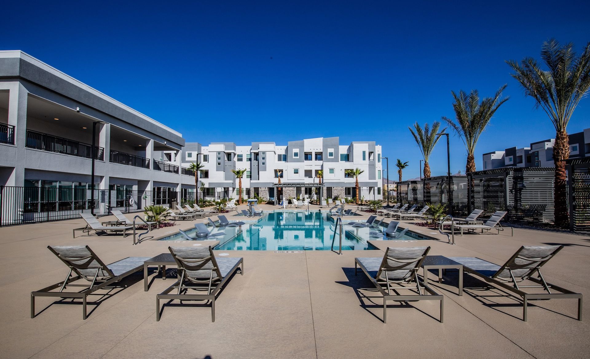 Pool area with lounge chairs, palm trees, and modern buildings under a blue sky.