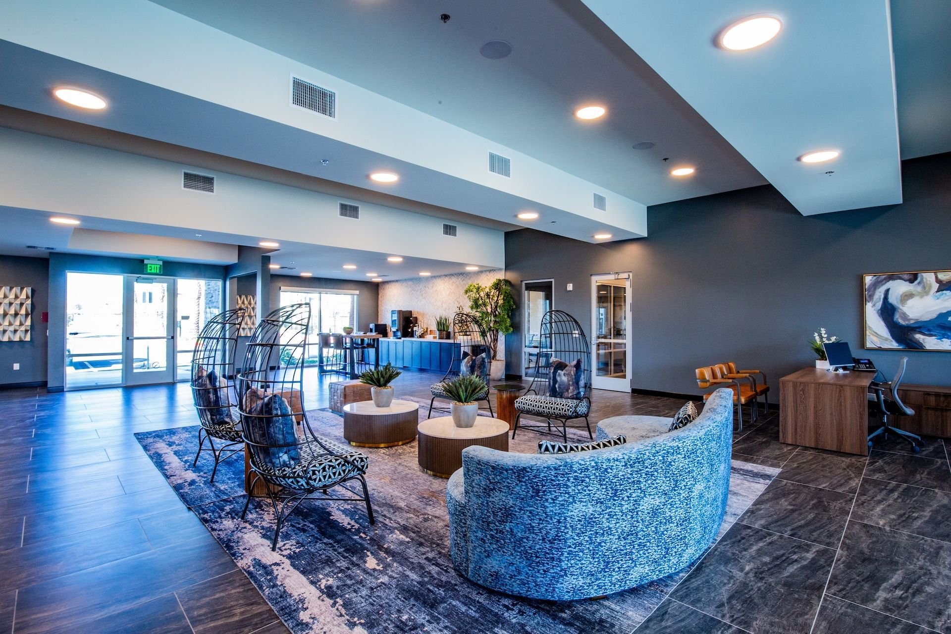 Lobby with seating, tables, and a front desk. Gray walls and flooring, with blue and white patterned furniture.