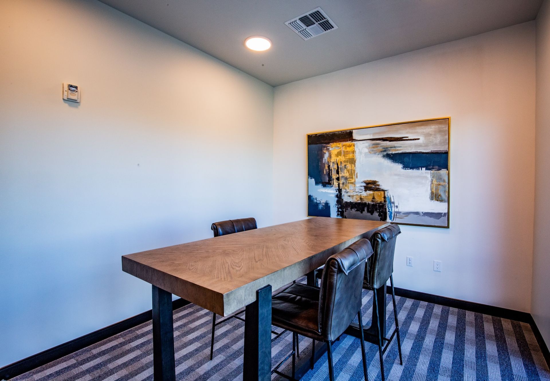 Small meeting room with a wooden table, artwork, and two chairs. Striped carpet and light walls.