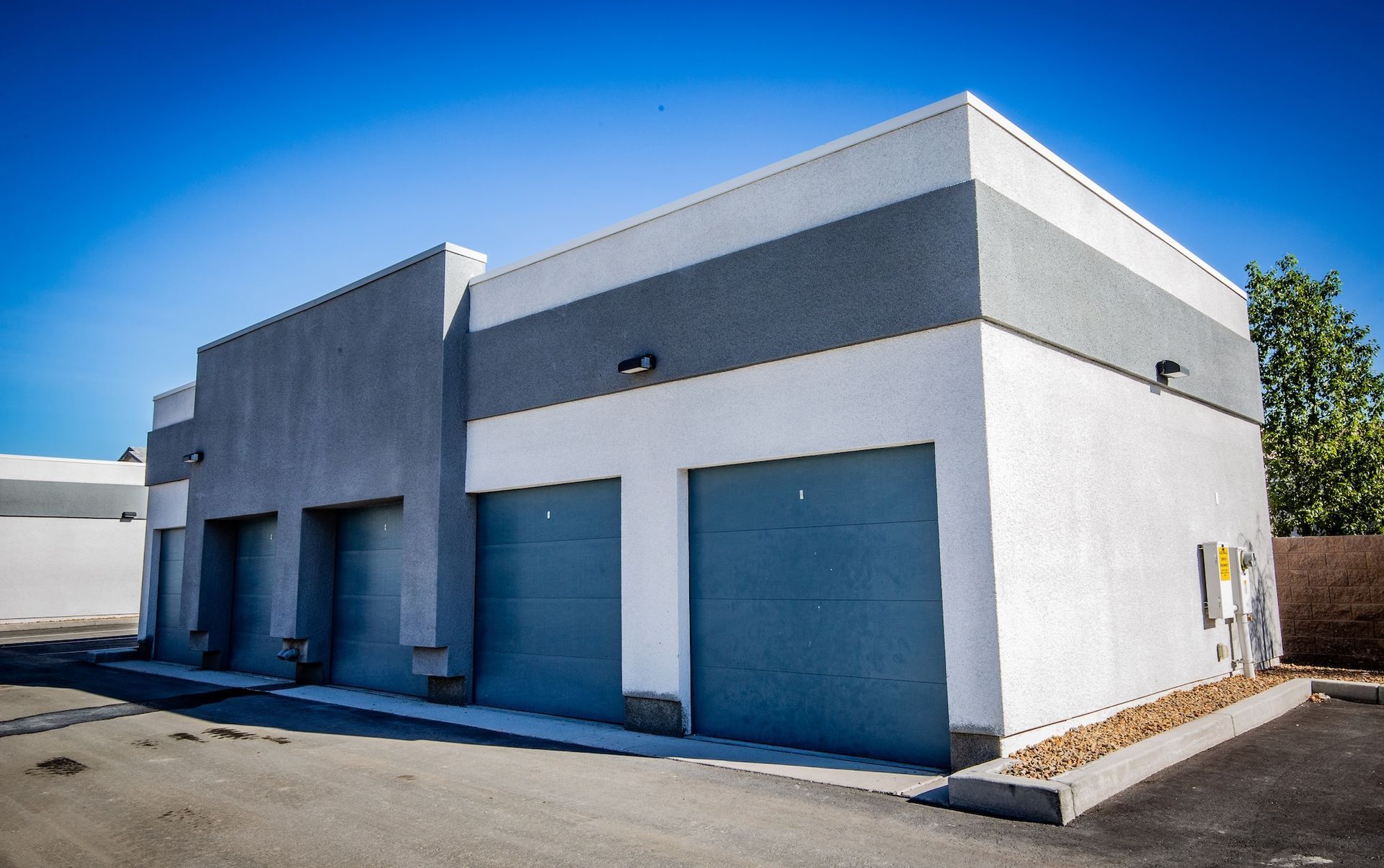 Three-bay industrial building, gray and white stucco, blue garage doors, bright blue sky.