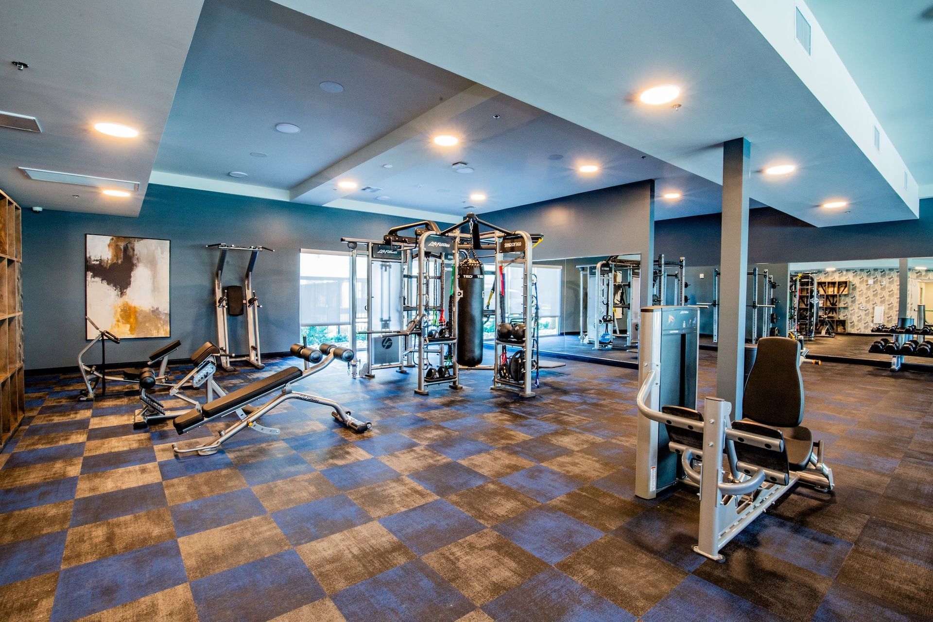 Gym interior with weight machines, benches, and a multi-station cable machine. Blue and gray floor tiles.