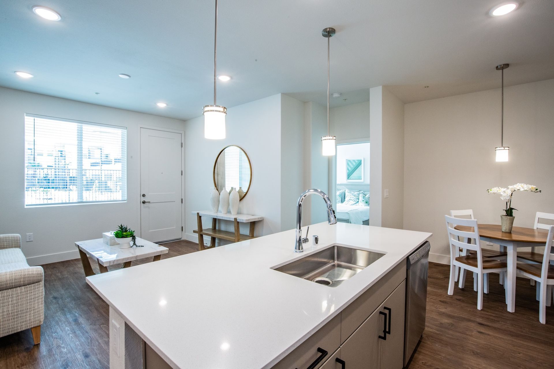 Modern kitchen with island, sink, and dining area. White cabinets, light wood table, and pendant lights.