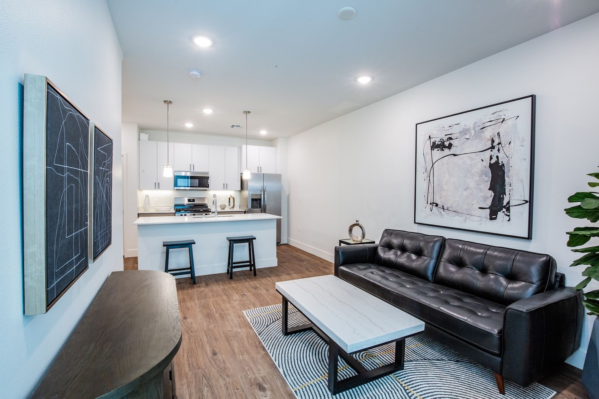 Modern apartment living room with black leather sofa, open kitchen, artwork, and hardwood floors.