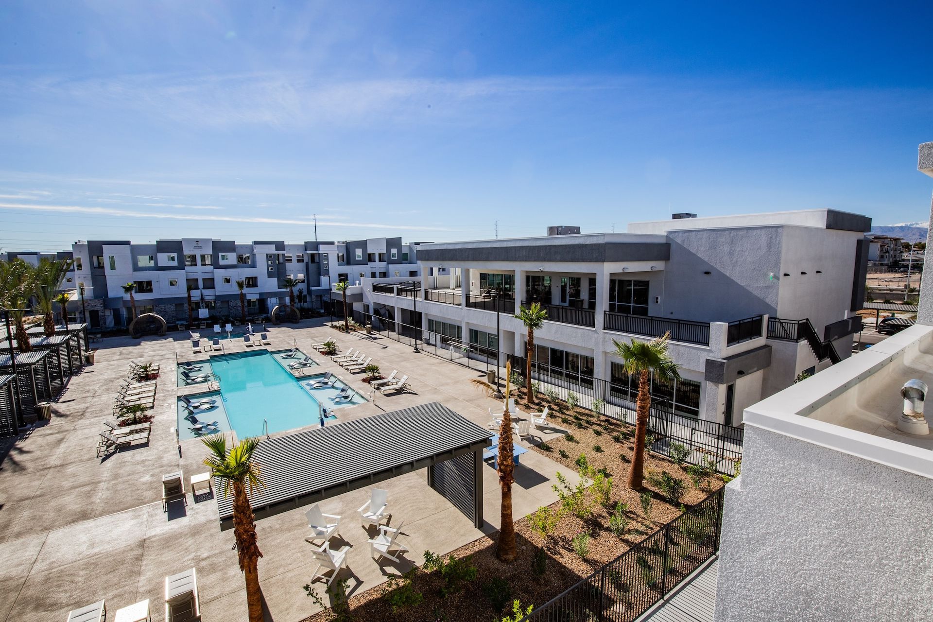 An aerial view of a modern apartment complex with a pool, palm trees, and clear blue sky.