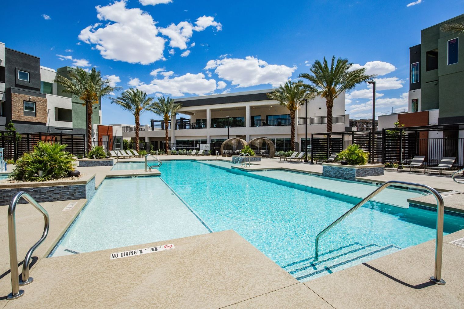 A large swimming pool is surrounded by palm trees and buildings.
