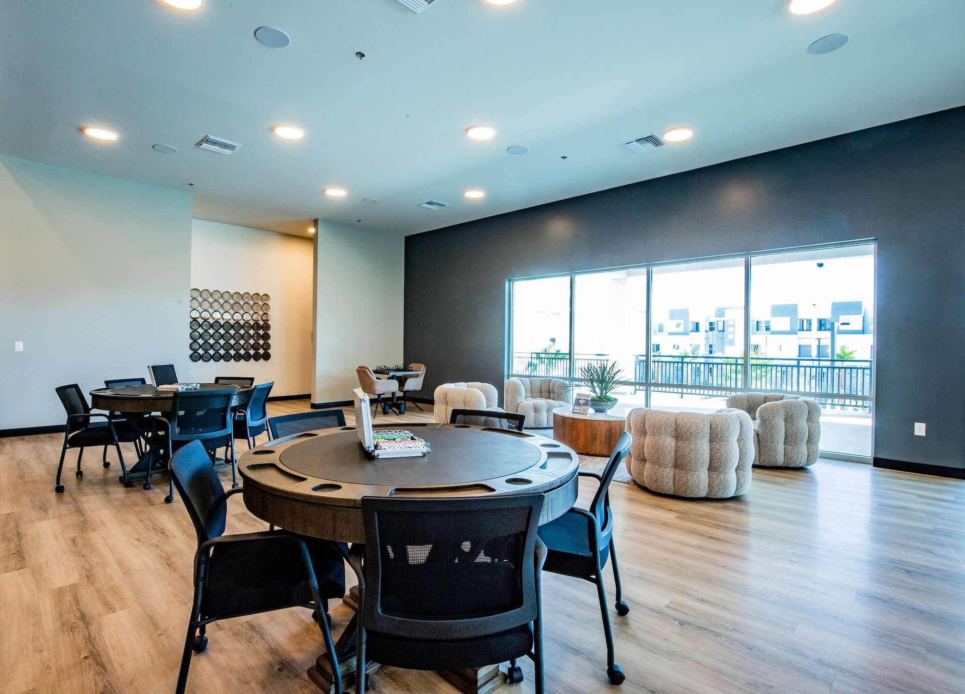 A community room with card tables, chairs, and window seating. Bright, neutral colors and overhead lighting.