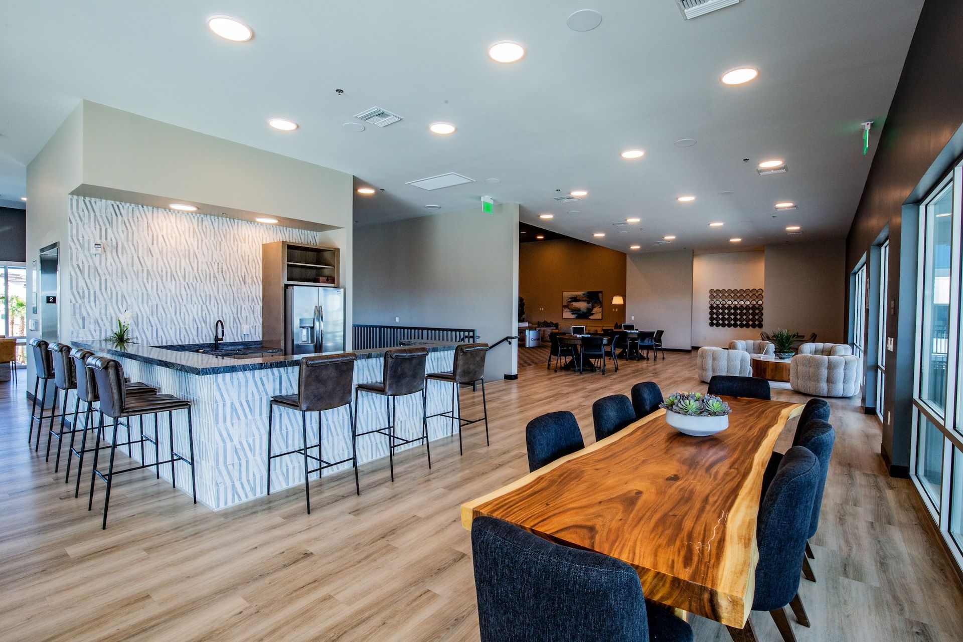 Lounge area with a long wooden table, bar with stools, and large windows. Beige walls and light wood floors.
