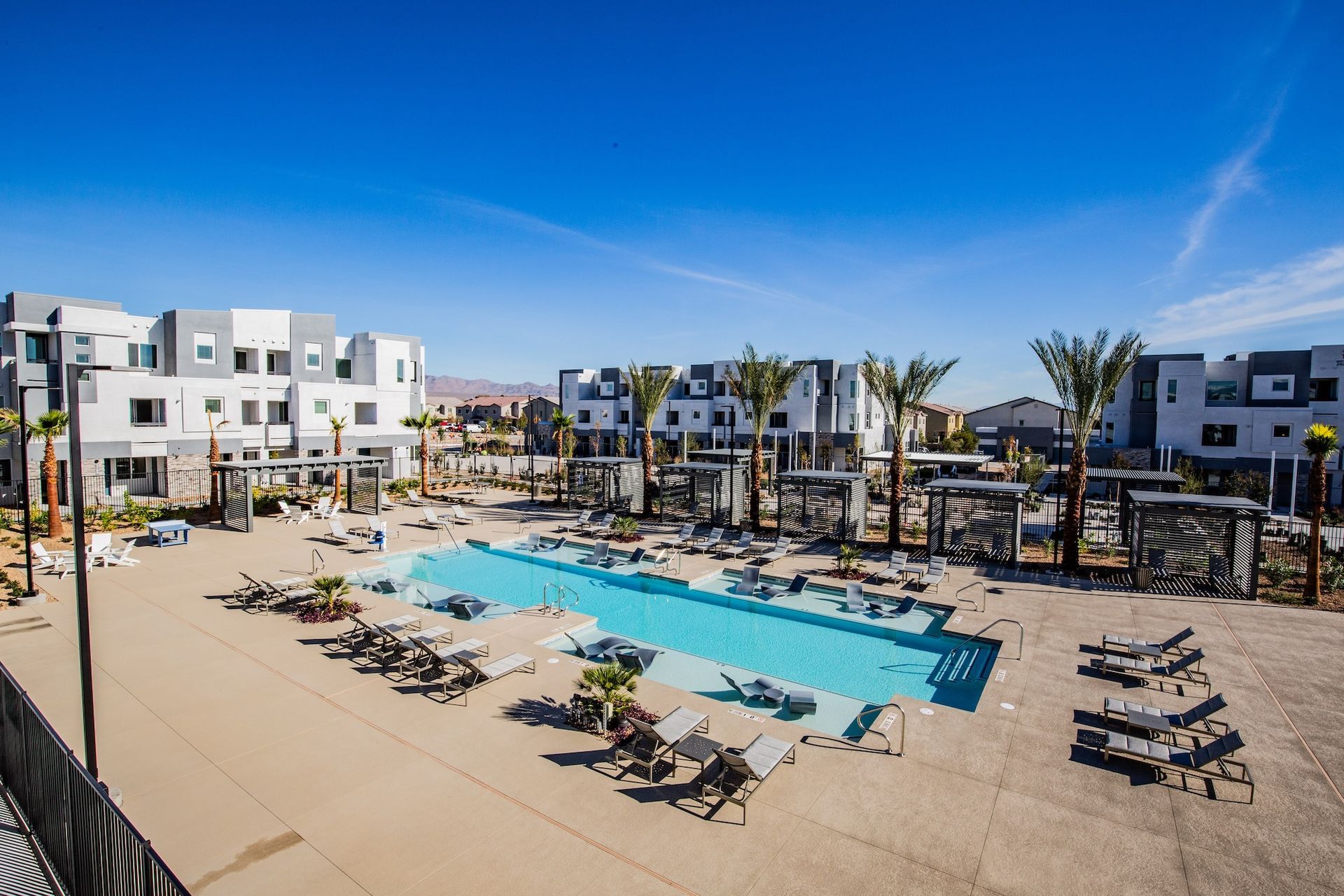 Pool and lounge area surrounded by modern townhouses under a bright blue sky.