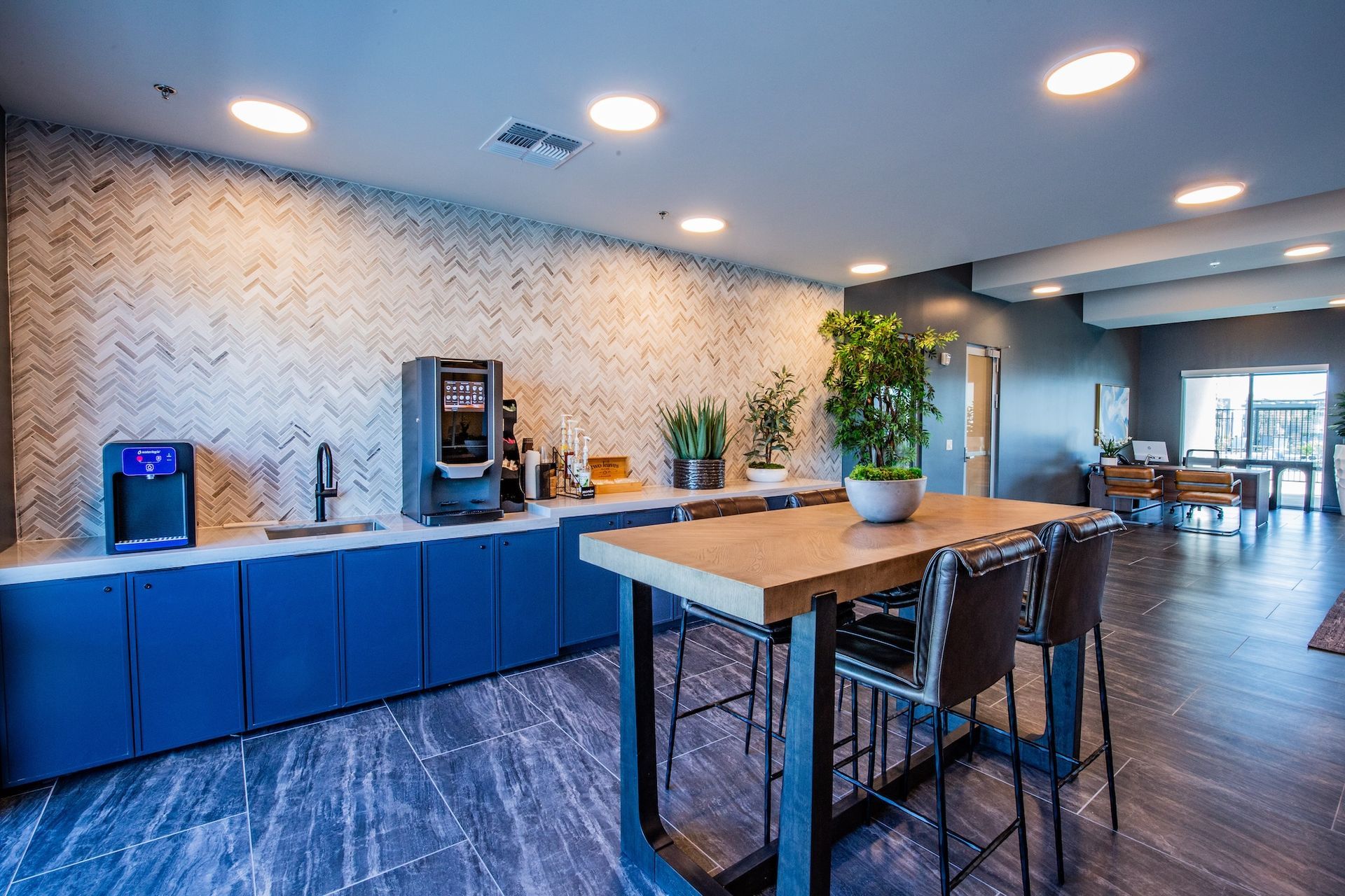 Modern break room with coffee station, blue cabinets, and a tall wooden table with stools.