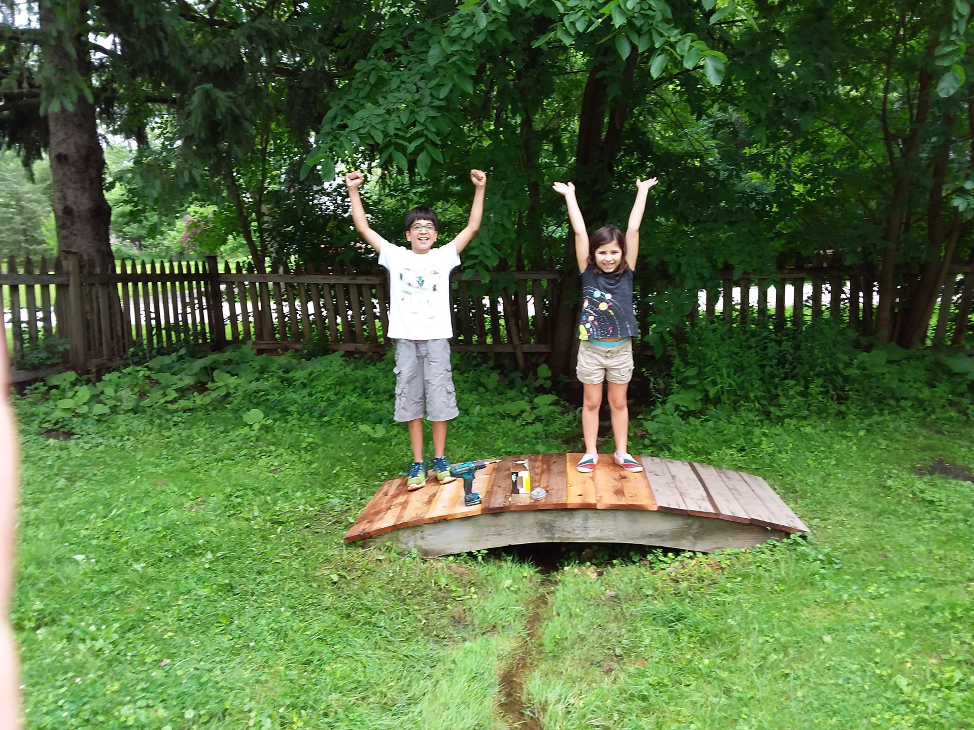 Two children with arms raised stand on a small wooden bridge over a shallow stream in a grassy yard.