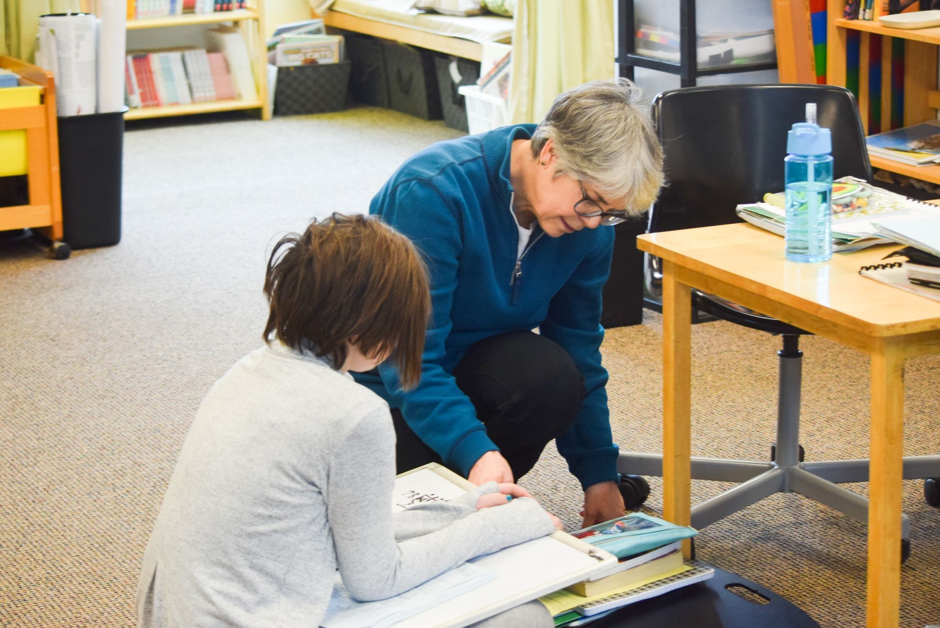 Woman helping another person on the floor with papers, in a room with shelves and a desk.