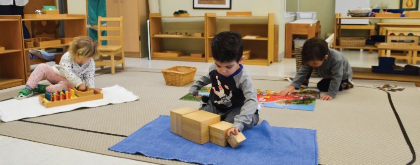 Three children playing with blocks and toys on the floor in a classroom.