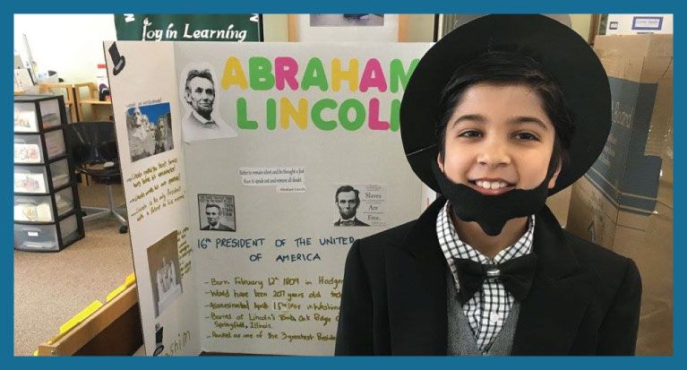 Young boy dressed as Abraham Lincoln, smiles in front of a school project.