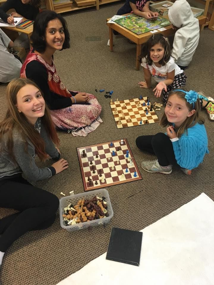Four children sit around two chess boards on the floor, smiling and playing.