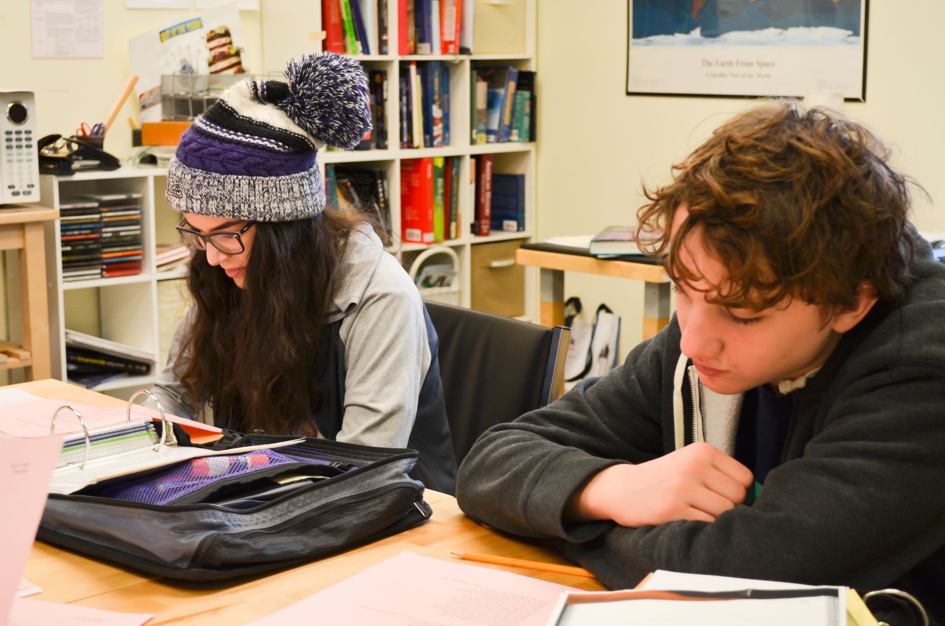 Two people studying at a table. One wears a hat, glasses, and jacket; the other, a hoodie. Bookshelves in the background.