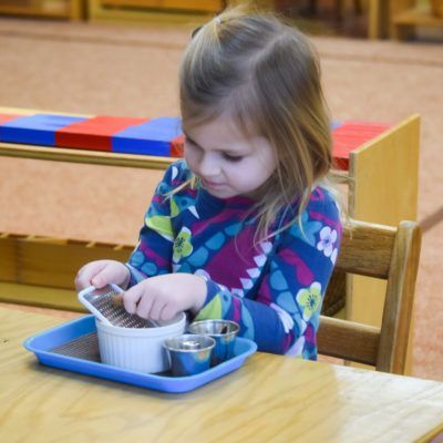 Child grating something into a bowl, seated at a table. Blue tray with bowl, metal cups. Wooden chair and table.