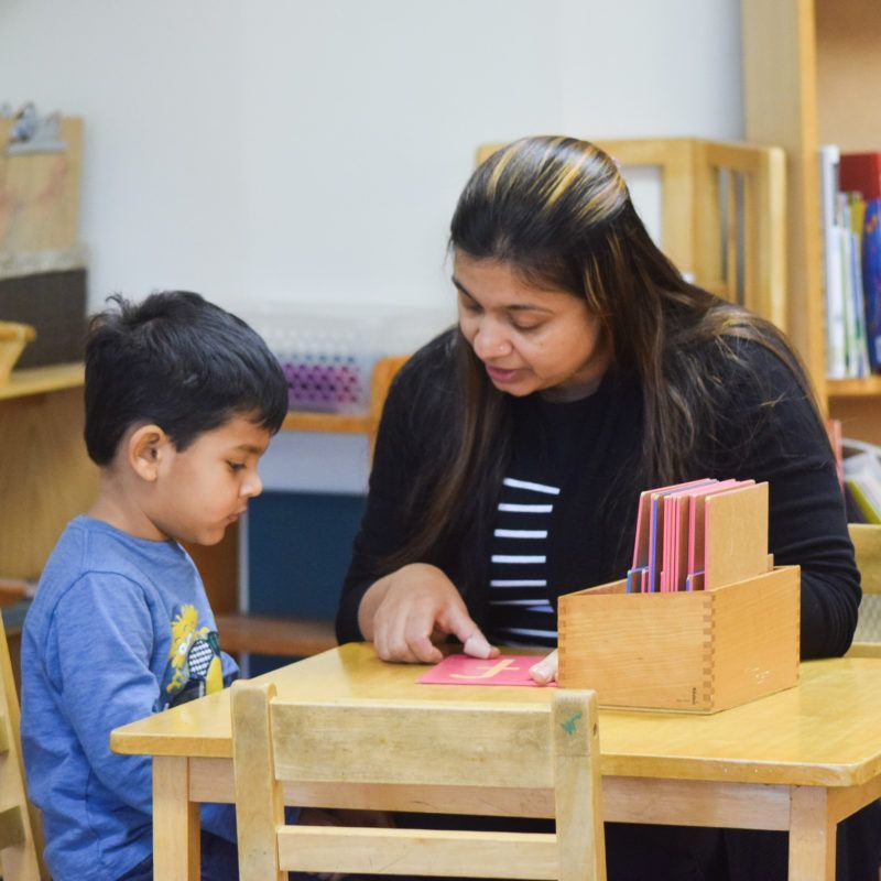A woman points at a card for a child at a wooden table in a classroom.