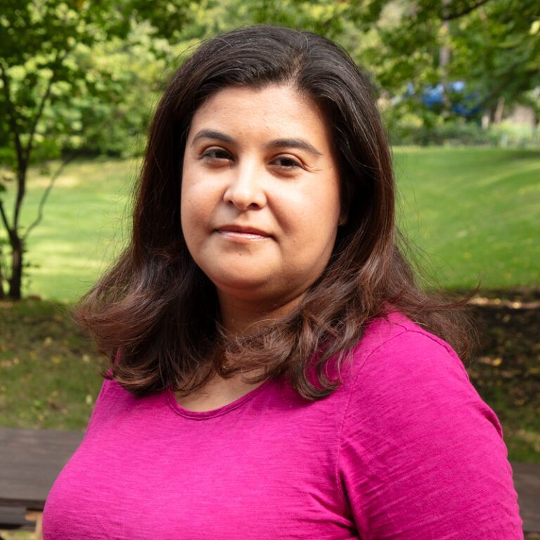 Woman in a pink shirt smiles outdoors, with trees and grass in the background.