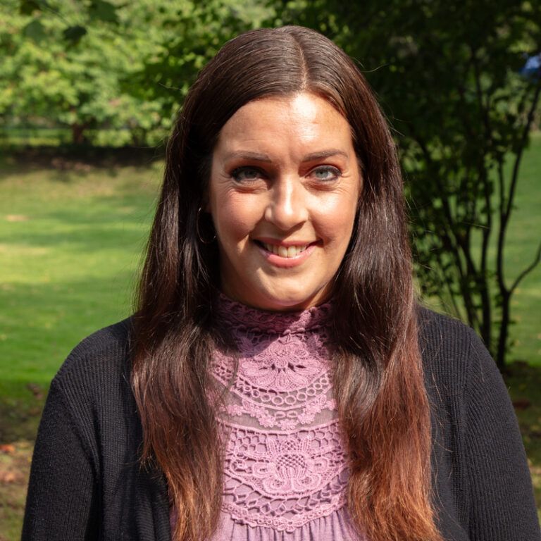 Woman with long brown hair, smiling, wearing a pink lace top and black cardigan, outdoors.