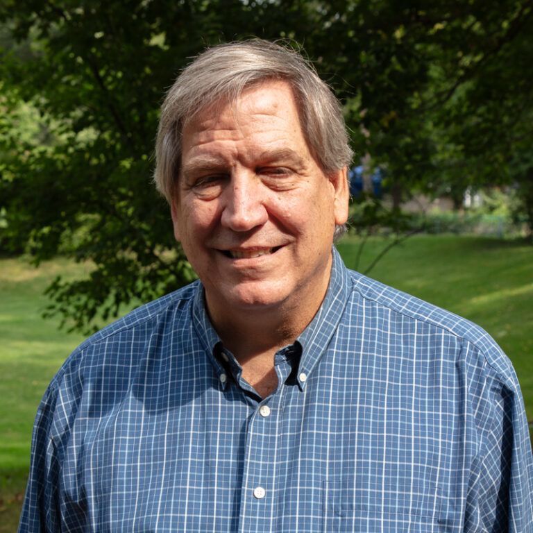 Man with graying hair, smiling, wearing a blue plaid shirt, outdoors in front of greenery.