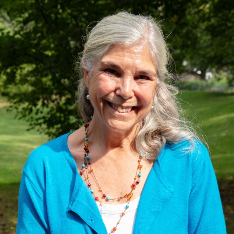 Woman with gray hair smiles, wearing a blue sweater and beaded necklace, outdoors.