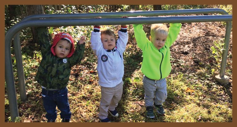 Three young children hanging from a metal bar in an outdoor setting.