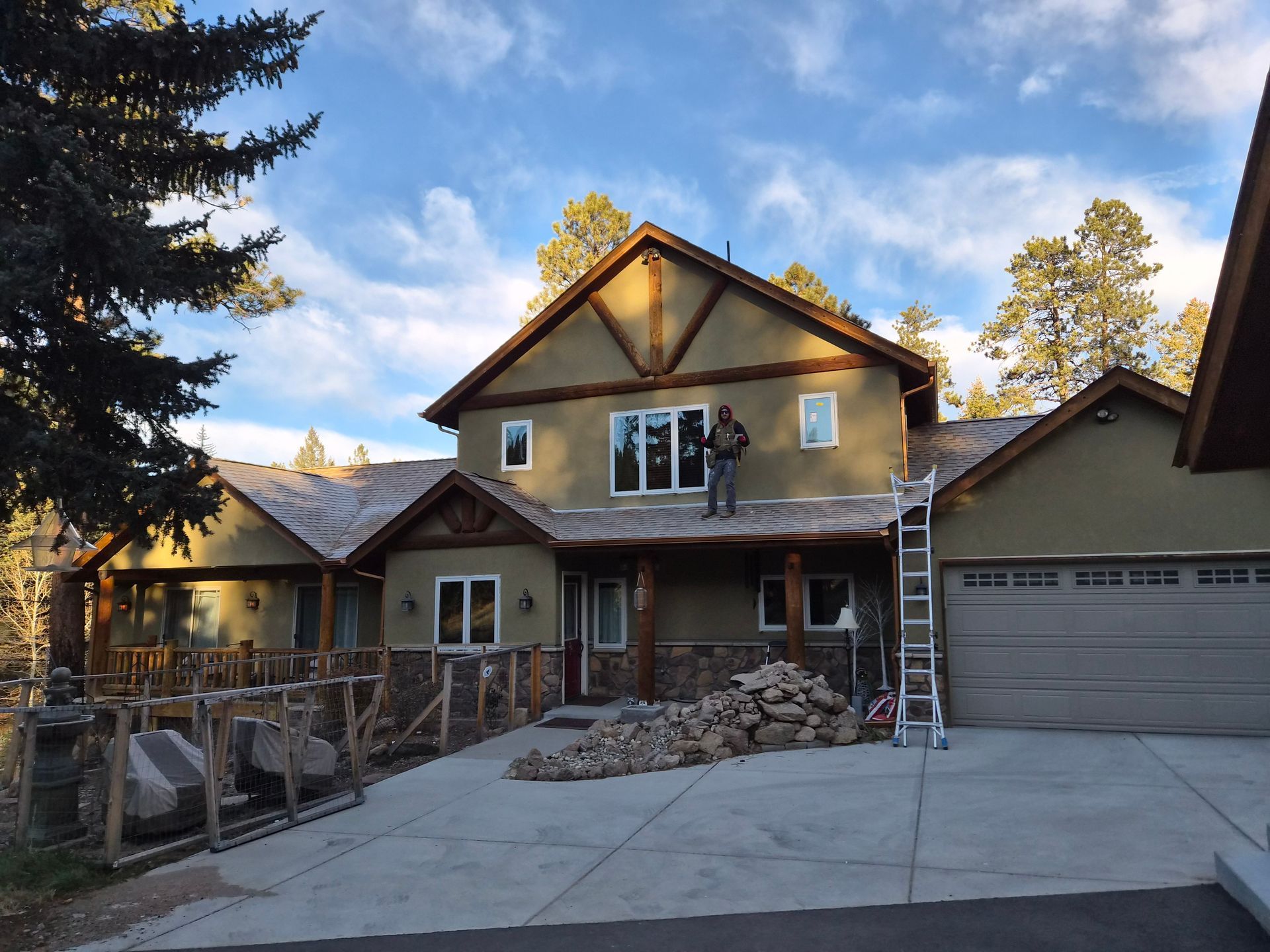 A person stands on the roof of a two-story home under construction, with building materials piled in the driveway.
