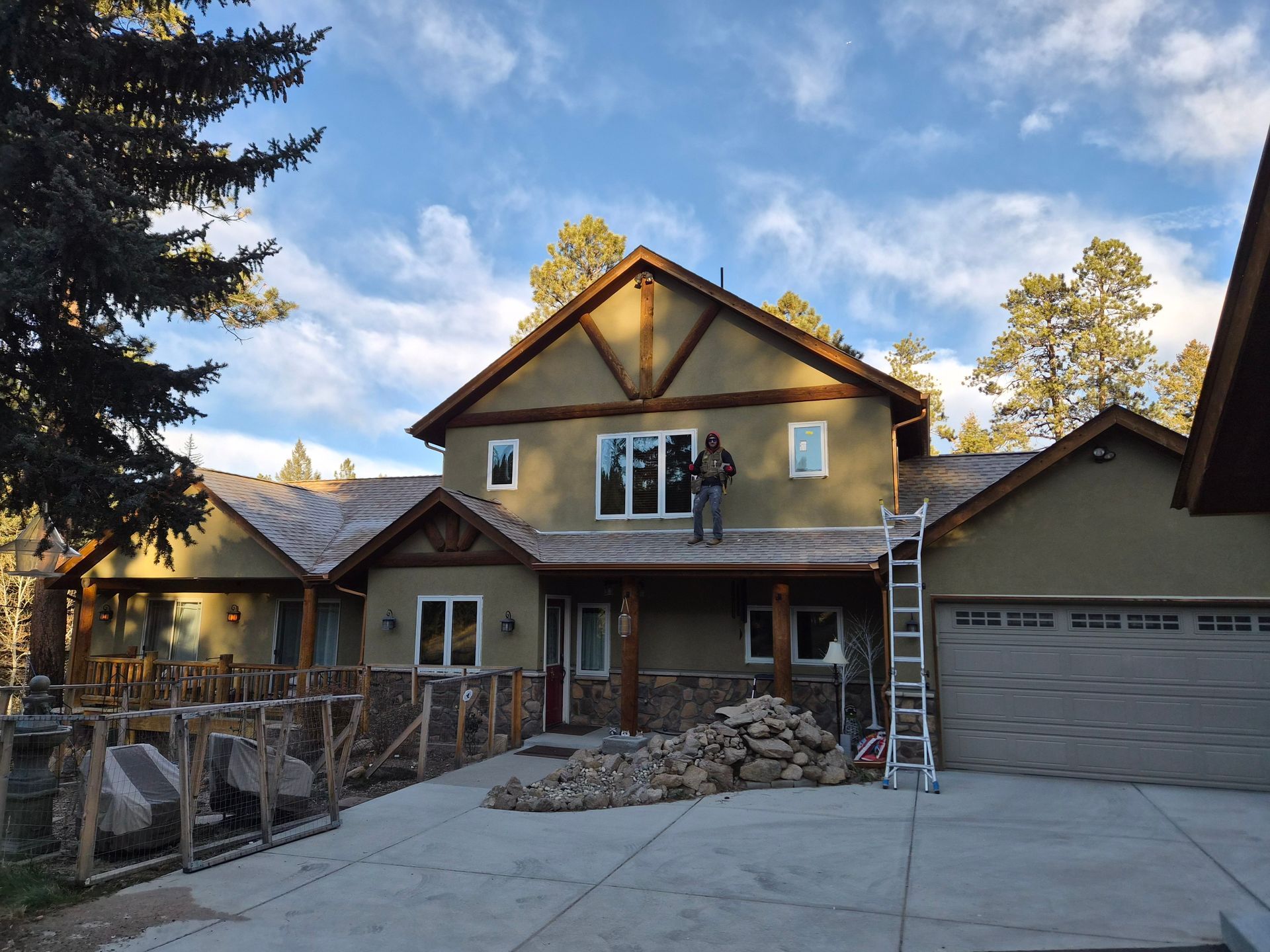 A person stands on the roof of a beige house with stone siding, near a ladder and a pile of rocks on the driveway.