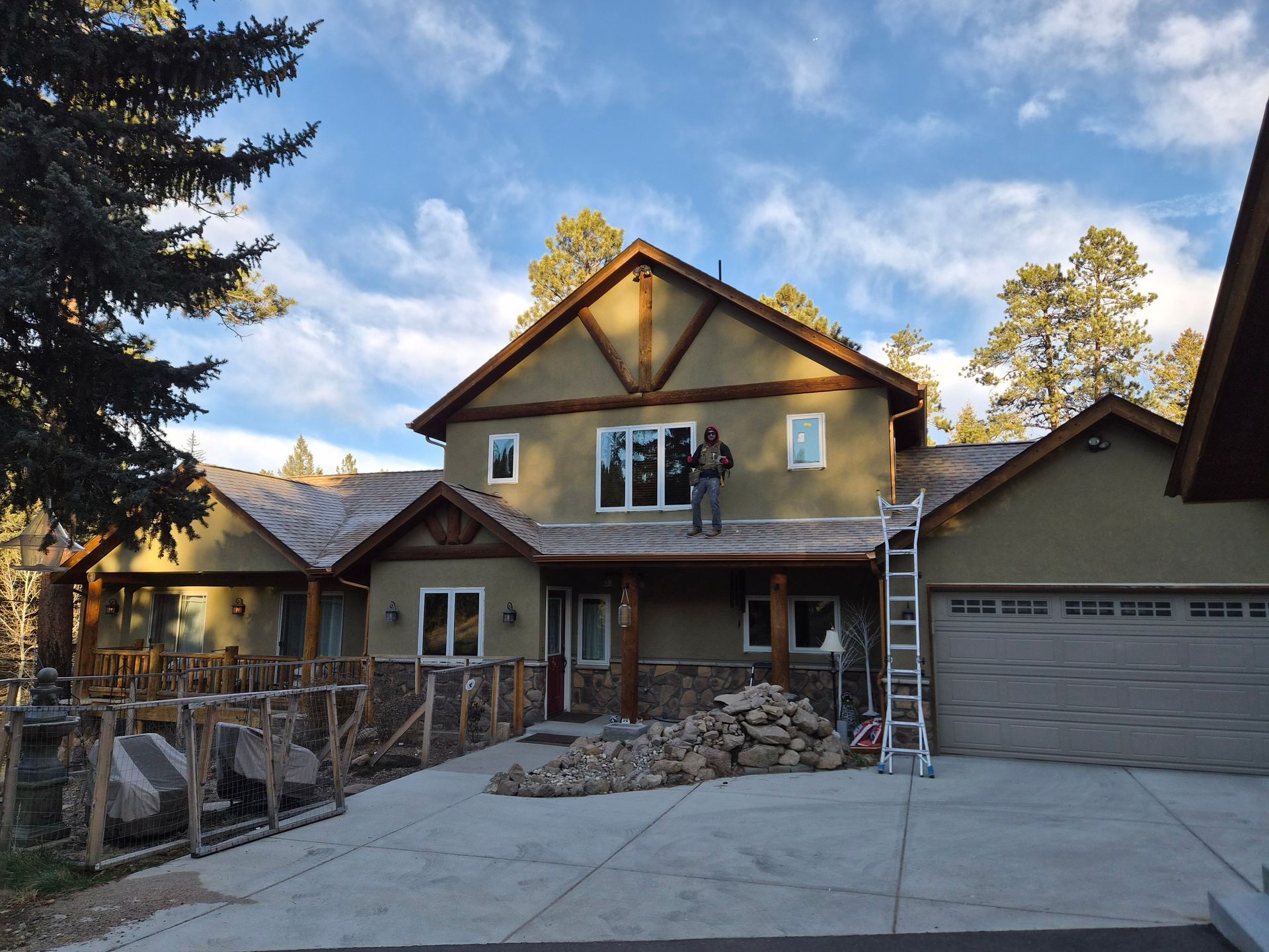 A person on a ladder repairs the roof of a two-story home with stucco siding and stone accents under a sunny blue sky.