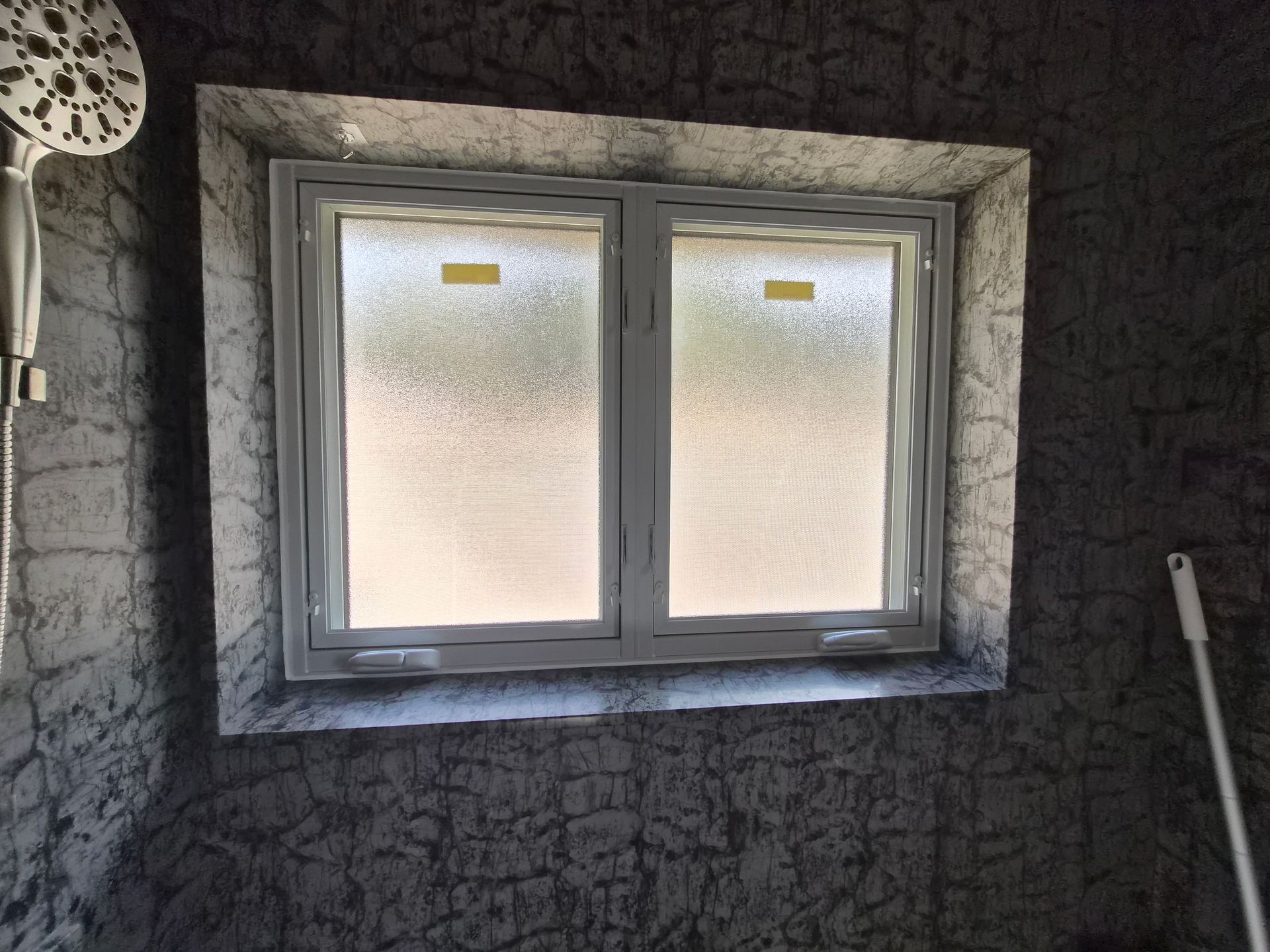 A white-framed window with frosted glass set into a dark, textured gray wall in a bathroom with a showerhead visible.