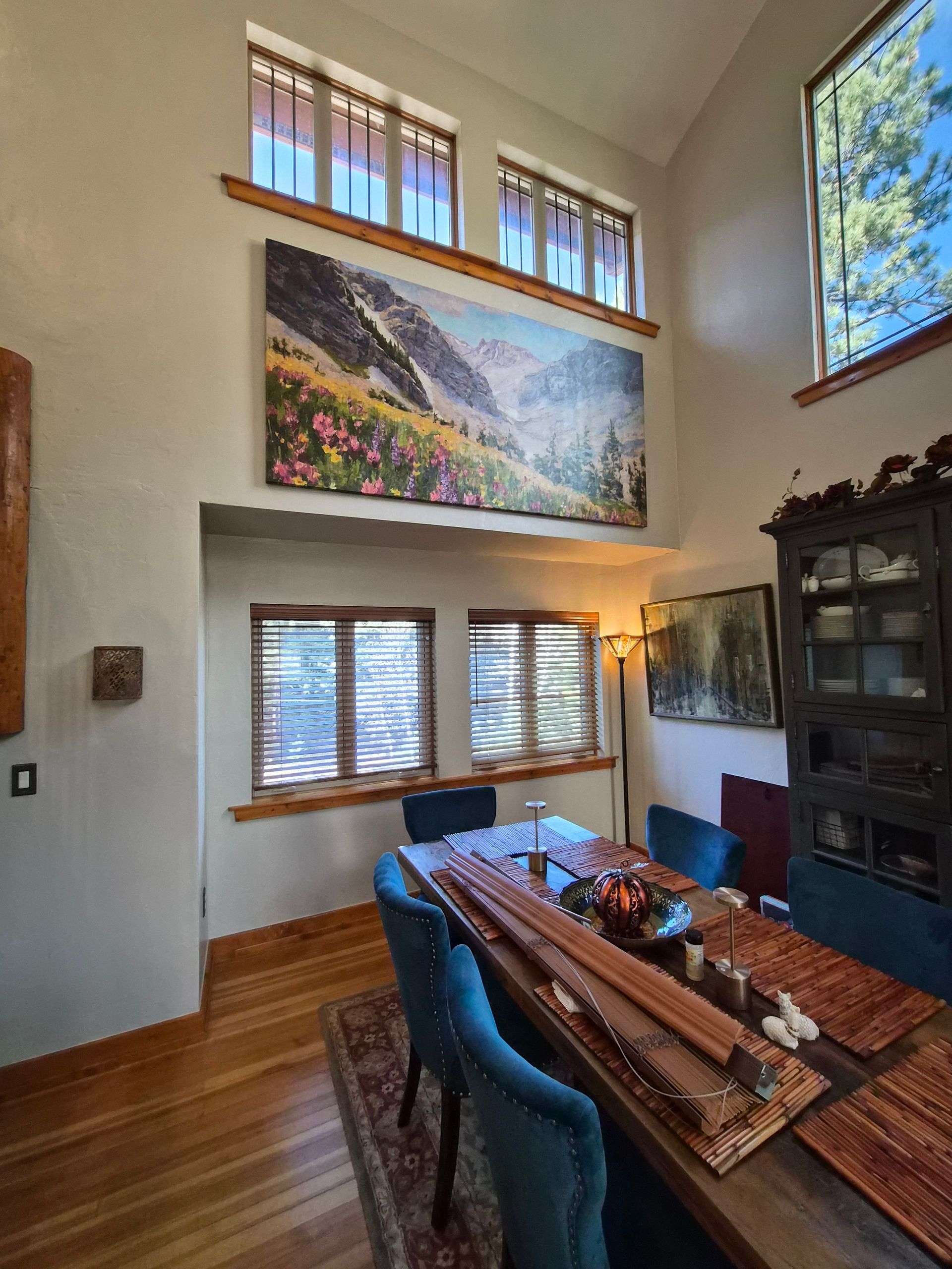 A dining room with a long table, blue chairs, a large landscape painting above a window, and wood-paneled walls.