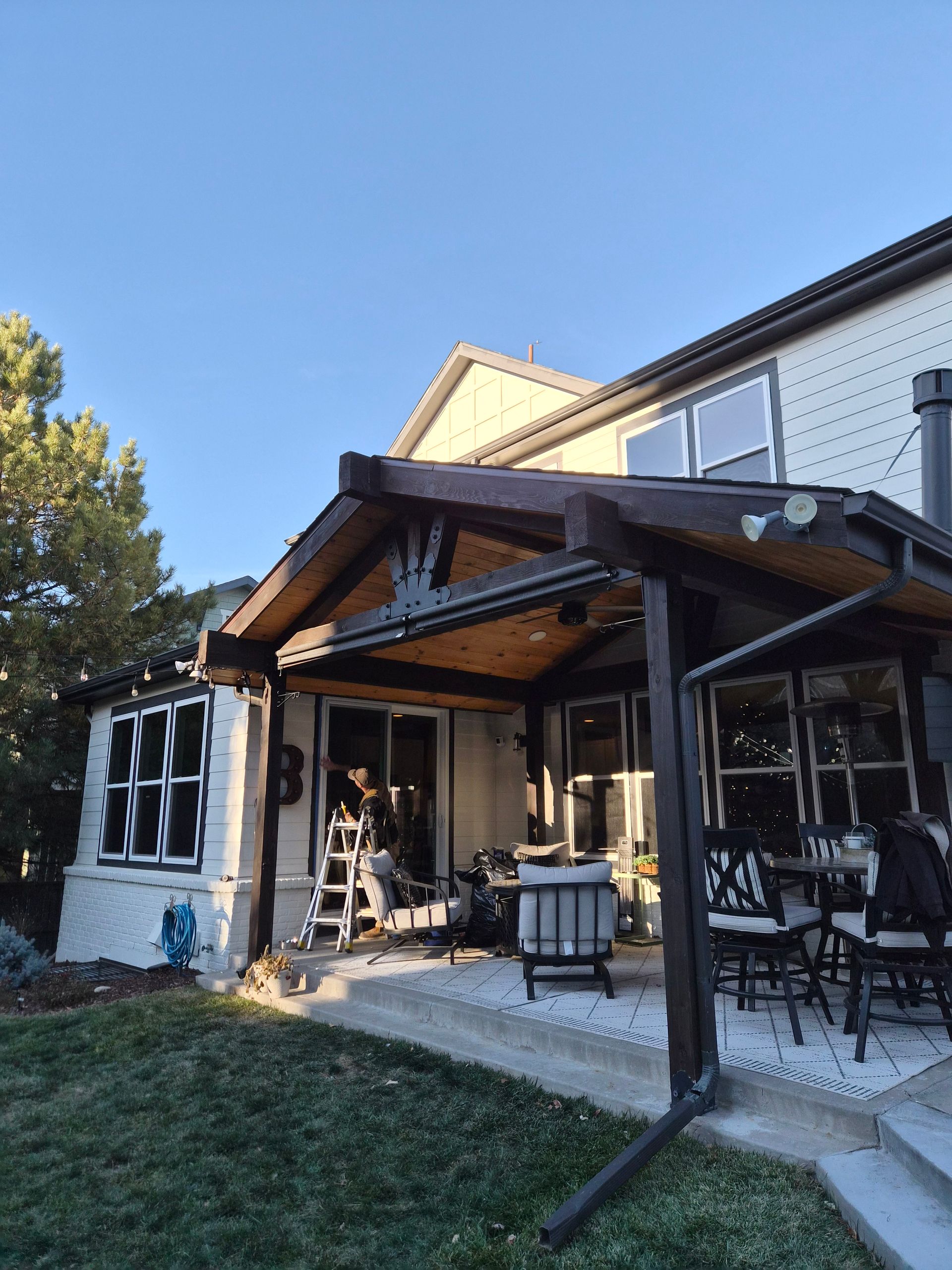 A person on a ladder working on the exterior of a home under a covered patio with outdoor furniture.